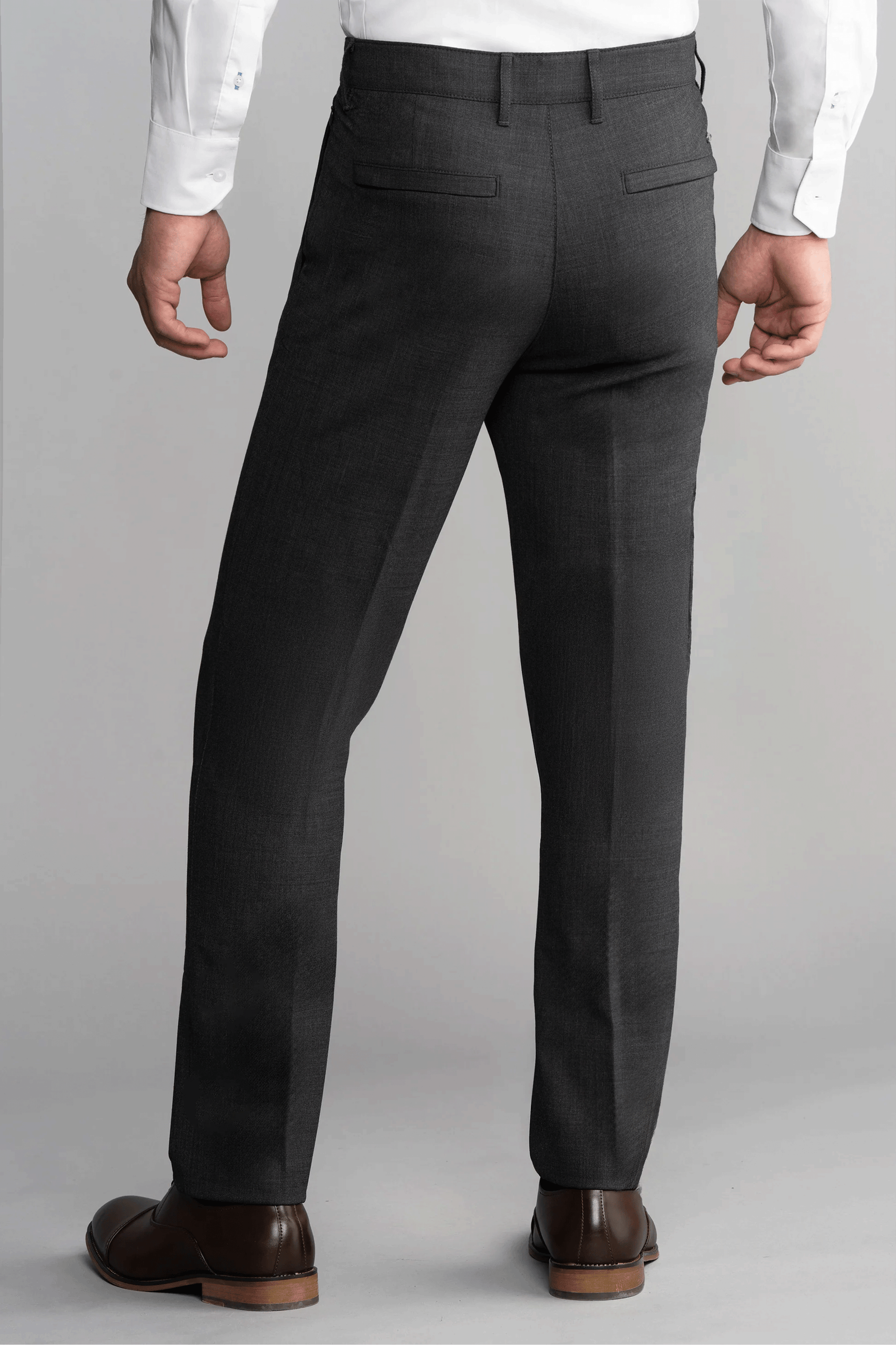 A man stands facing away, wearing Twillory Hybrid Wool Pants in Grey Heather, a white long-sleeve shirt, and brown dress shoes. The pants feature two back welt pockets. A plain light gray background highlights the formal look.