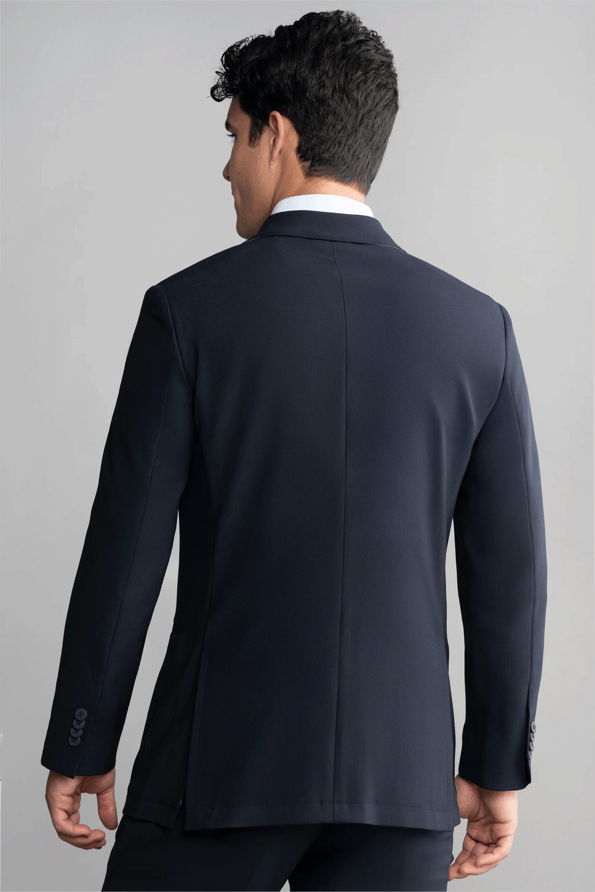 A man with short, dark, curly hair is seen from behind in a Twillory Hybrid Wool Blazer // Navy, featuring sleeve buttons. He stands against a plain light gray background with his head turned slightly left.