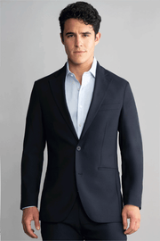 A man with short, curly dark hair models the Twillory Hybrid Wool Blazer in Navy over a light blue dress shirt. He stands facing forward with a neutral expression and relaxed hands against a plain light gray background.
