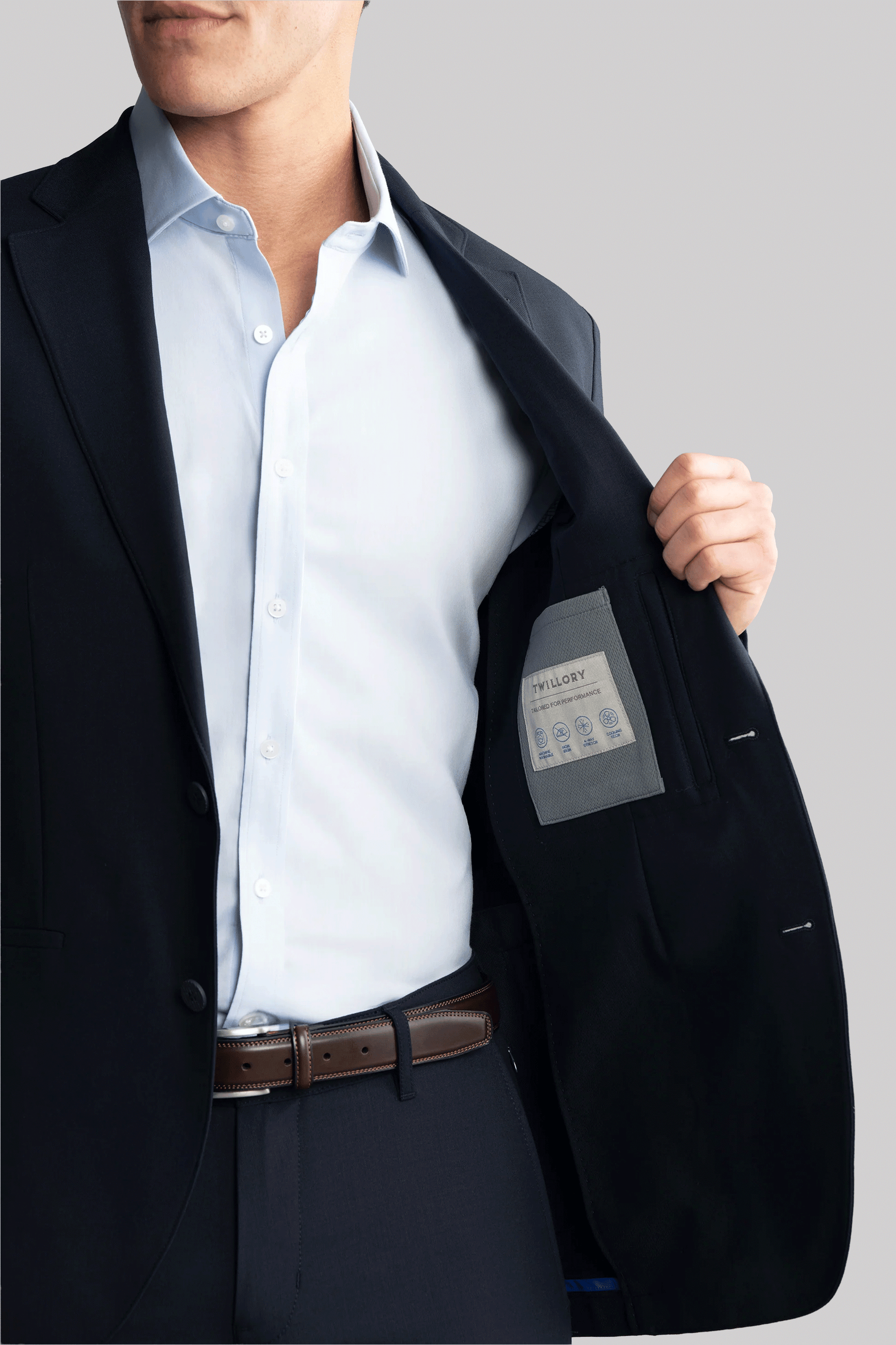 A man in a navy Twillory Hybrid Wool Blazer opens the jacket to reveal its label, highlighting 4-way stretch and Hybrid Wool. He wears a light blue shirt, with only the lower half of his face visible against a light gray background.