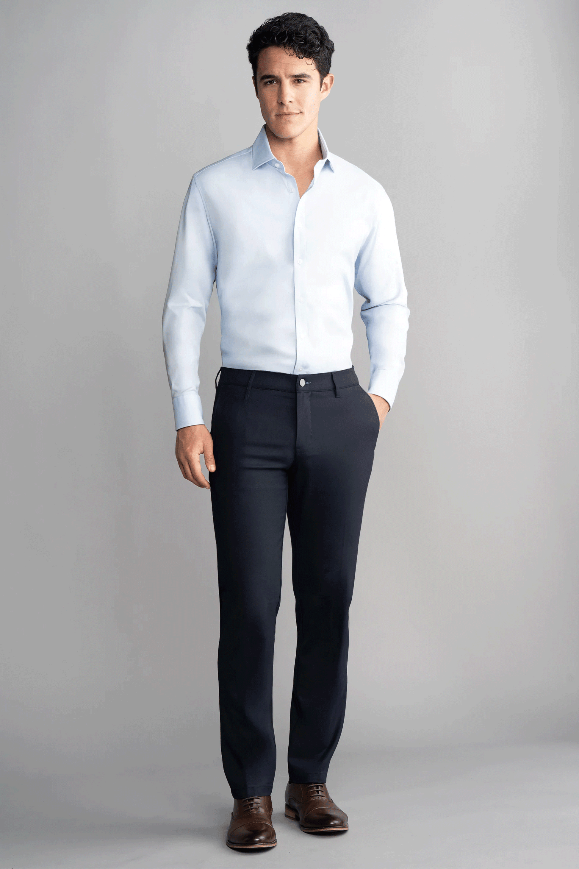A man with short dark hair and a neutral expression wears a light blue dress shirt tucked into Twillory's Hybrid Wool Pants // Navy, featuring 4-way stretch. He stands against a gray background with one hand in his pocket and brown leather dress shoes.