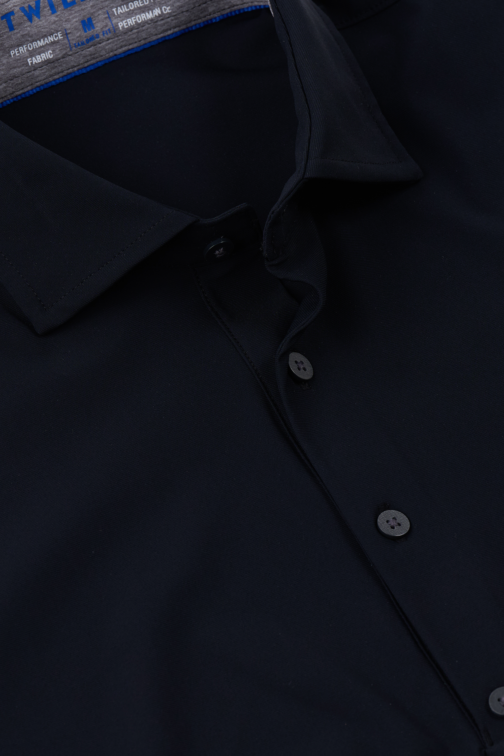 Close-up of the Twillory Event Polo Solid in black, featuring a classic collar and black buttons. The performance fabric looks smooth and tailored, with blue text partially visible on the inner label inside the collar.