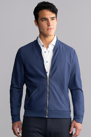 A man with short dark hair wears the Twillory AIR Bomber in blue over a white button-down shirt. He stands against a plain gray background, looking to the side with a neutral expression and his hands relaxed by his sides.