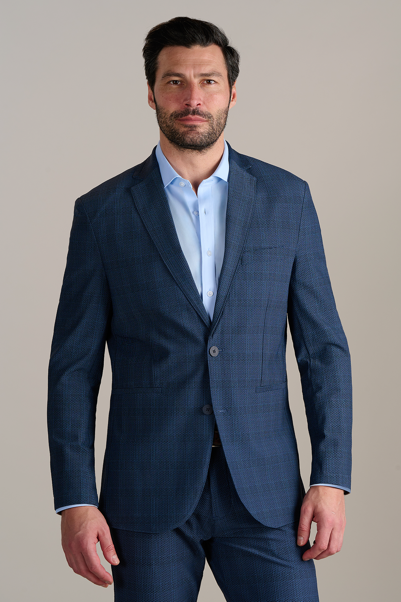 Man wearing a blue checkered suit with a light blue dress shirt, standing in front of a neutral background.
