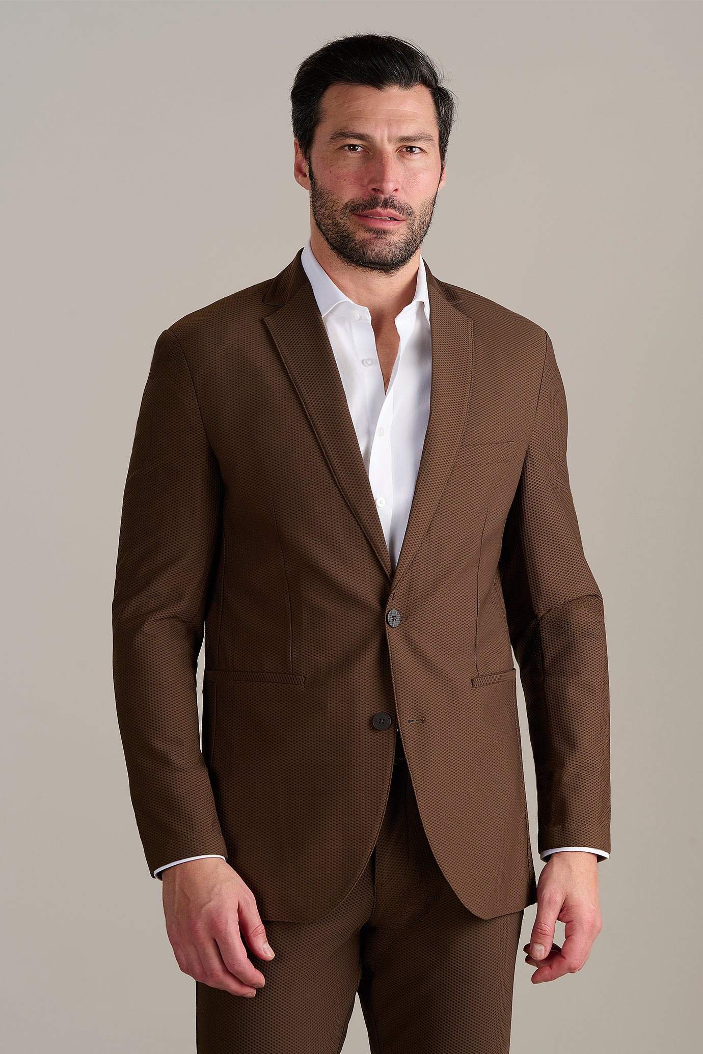 Man wearing a brown suit with a white dress shirt, standing against a neutral background.
