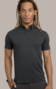 A man with long, light brown dreadlocks wears the Twillory Short Sleeve Polo Dot in Charcoal, featuring a dress collar and small dotted pattern. He stands facing forward against a beige background, showing his smartwatch and neutral expression.