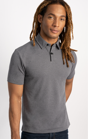 A person with long, light brown dreadlocks stands against a white background, wearing the Twillory Short Sleeve Polo Contrast in Grey Black. The shirt features a buttoned collar and cooling tech. They have a relaxed expression and look at the camera.