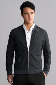 A young man with short curly dark hair wears the Twillory AIR Bomber // Heather Charcoal over a white dress shirt with blue buttons, standing against a plain light grey background and looking slightly left with a neutral expression.