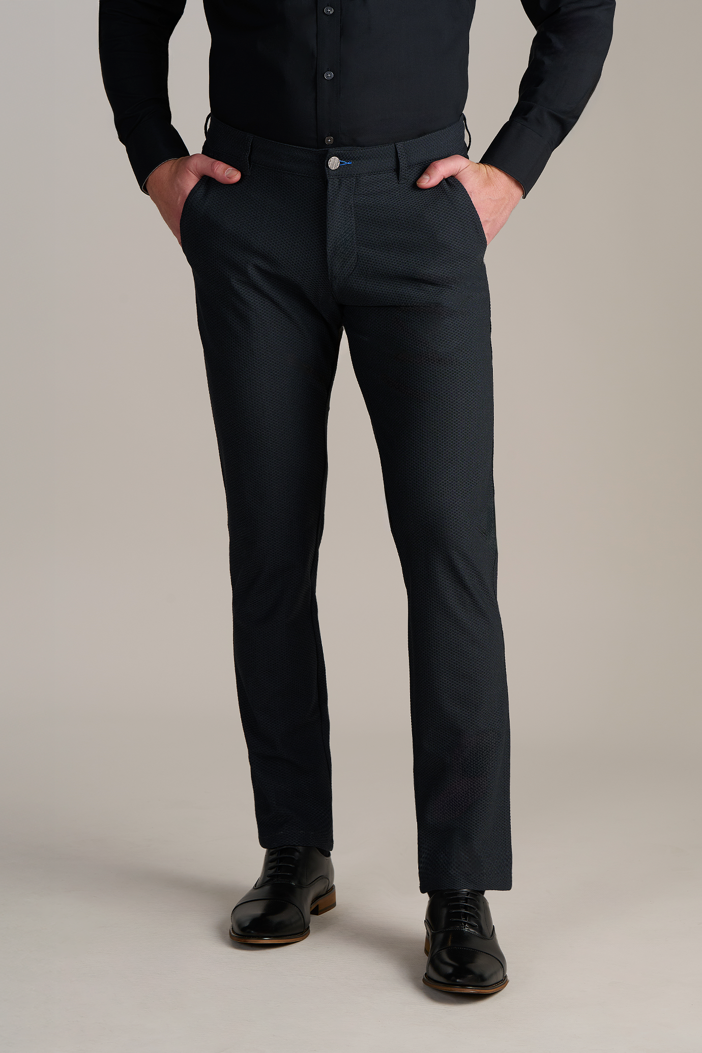 Man wearing black formal pants and shoes with hands in pockets against plain background.