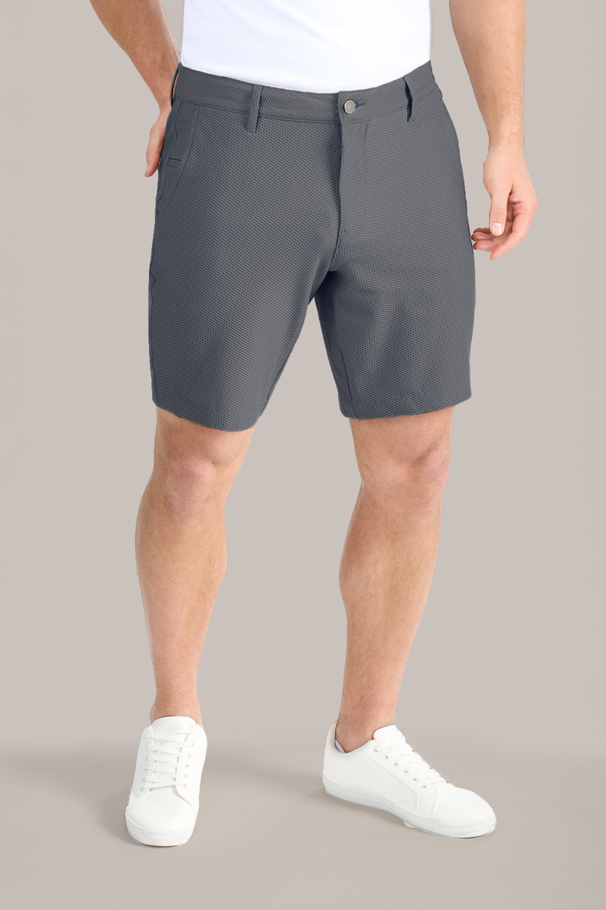 A man stands against a plain light background, wearing Twillory AIR Shorts made from 360° Airflow Fabric in gray, a white T-shirt, and white low-top sneakers. His left hand is in his pocket; his face is not visible.