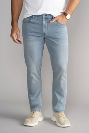 A man, visible from the shoulders down against a plain gray background, wears Twillory's Performance Denim // LIGHT WASH BLUE jeans in a tapered fit with one hand in his pocket, paired with a white T-shirt, beige sneakers, and a brown wristwatch.