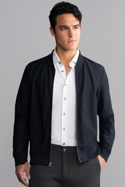A man with short dark hair and a thoughtful expression looks to the side against a gray background, wearing Twillory’s AIR Bomber // Navy over a white dress shirt with blue buttons and dark trousers.