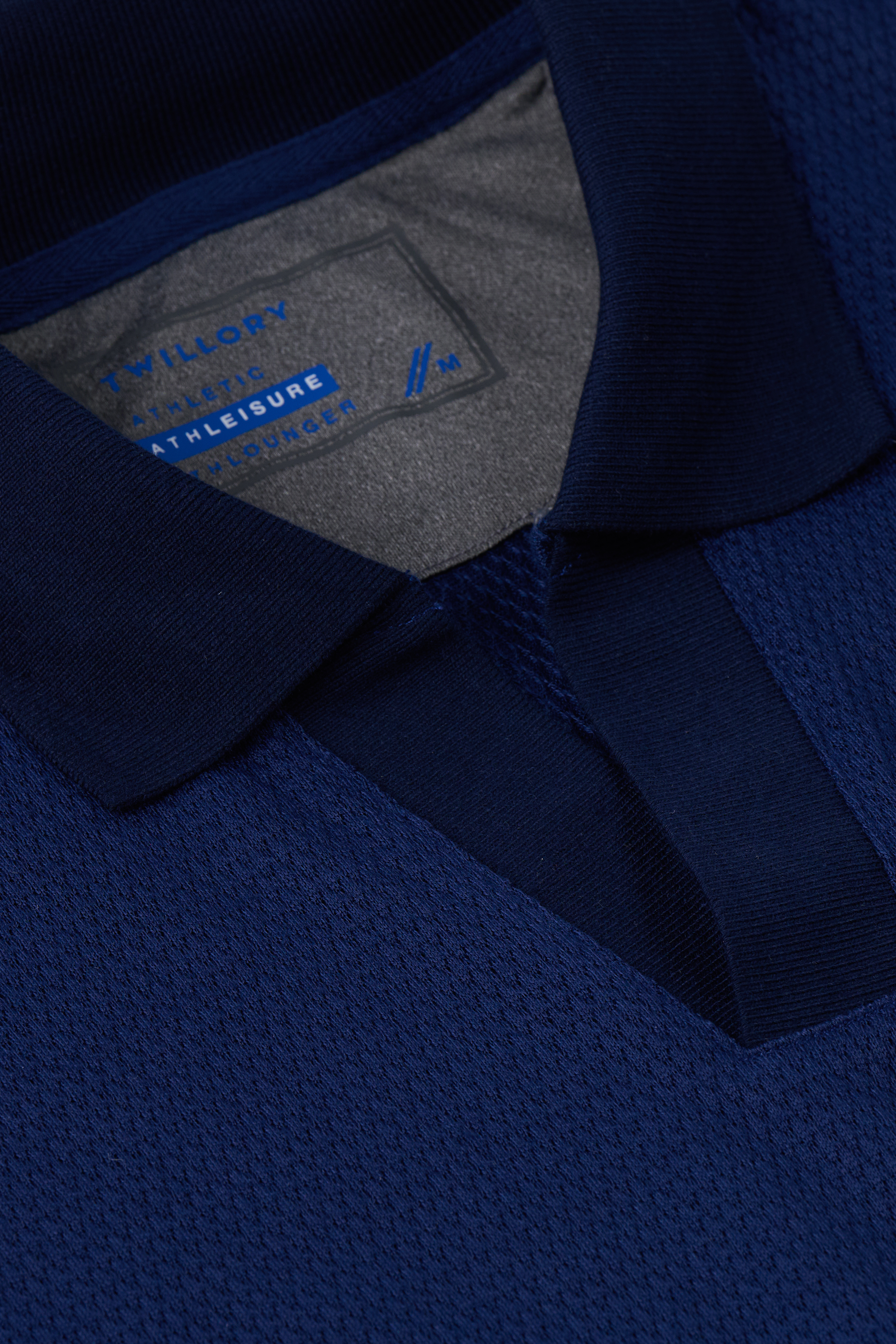 Close-up of the Twillory Short Sleeve Sweater Polo Solid in navy, featuring textured performance knit fabric and a short placket with hidden buttons. The collar label reads “ATHLEISURE” and size “M.” Wrinkle-free for easy wear.