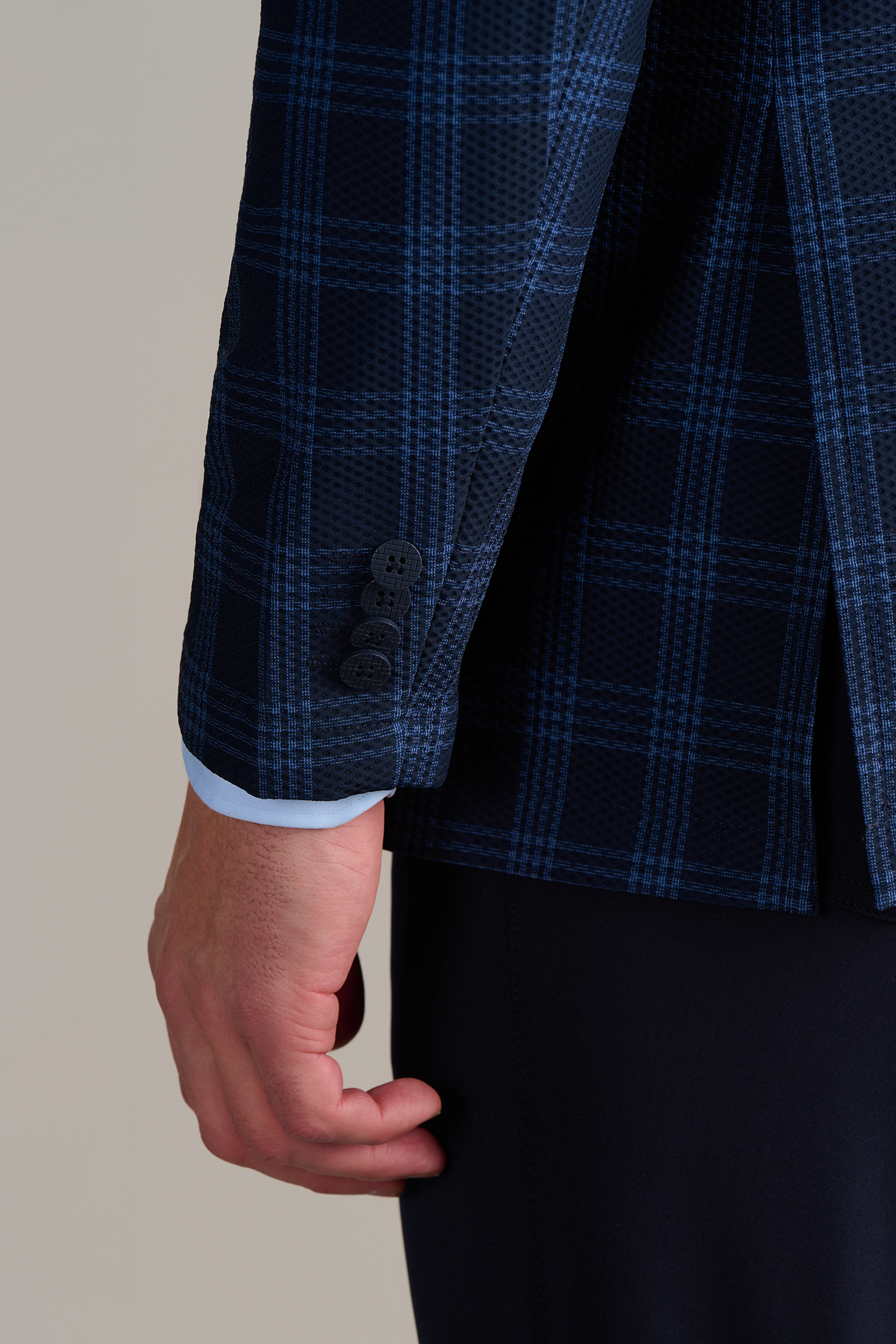 Close-up of a person's hand wearing a dark blue plaid suit jacket with blue buttons over a light blue shirt.