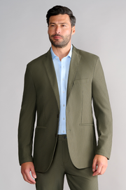 A man with short dark hair and a trimmed beard wears the Twillory AIR Blazer in Olive over a light blue dress shirt, standing against a plain light gray background. He looks slightly left with a neutral expression, his right hand relaxed by his side.