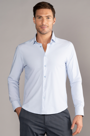 A man with short brown hair and light stubble stands against a plain gray background, wearing Twillory’s The Event Shirt // Powder Blue and gray trousers. He smiles slightly, with one hand in his pocket and the other relaxed by his side.