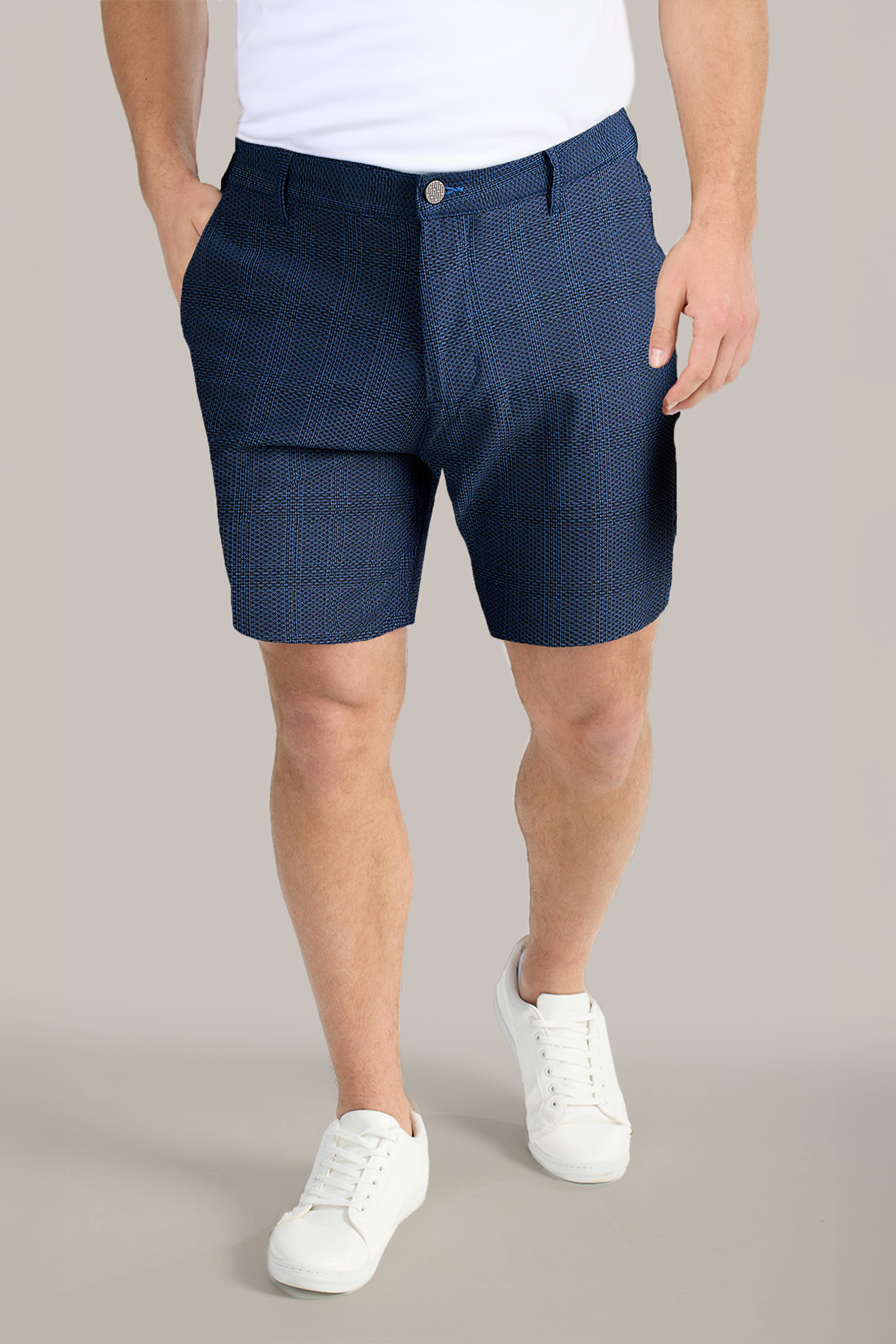 A person stands with hands in the pockets of Twillory AIR Shorts in dark blue pattern, paired with a white T-shirt and white sneakers. The plain background highlights this casual summer outfit. The face is not visible.