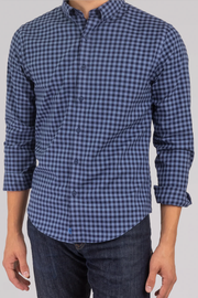 A person wears Twillory's untuck(able) Midnite Gingham—a smart casual, long-sleeve blue and black checkered shirt with rolled sleeves—paired with dark blue jeans. The image shows the torso and upper legs on a light background.