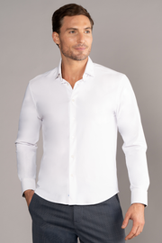 A man with short brown hair and light stubble wears Twillory's The Event Shirt // White, wrinkle-free with a relaxed fit and open collar, standing against a gray background—an ideal choice for modern men's officewear.