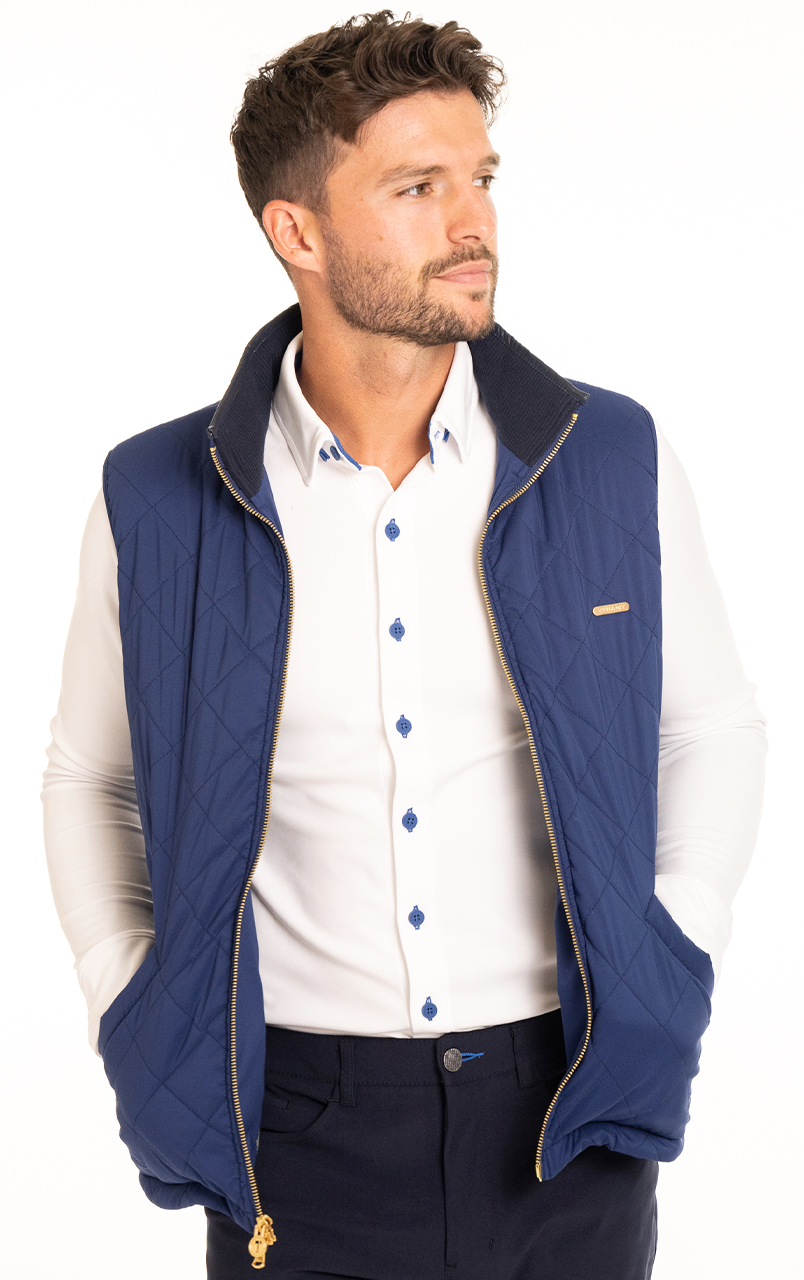 Man wearing a stylish blue quilted vest over a white shirt with blue accents, looking to the side, hands in pockets, on a white background.