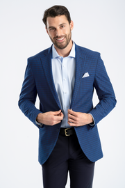 A man with short brown hair and a beard smiles as he buttons a Twillory Performance Blazer // Blue Check over a light blue shirt, styled with dark trousers, a black belt with gold buckle, and a white pocket square against a plain white background.