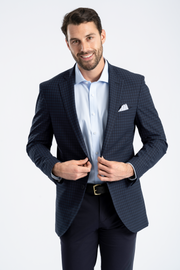 A man with short brown hair and a beard smiles while buttoning the Twillory Performance Blazer // Navy Check over a light blue shirt, paired with dark trousers, a black belt with gold buckle, and white pocket square against a white background.
